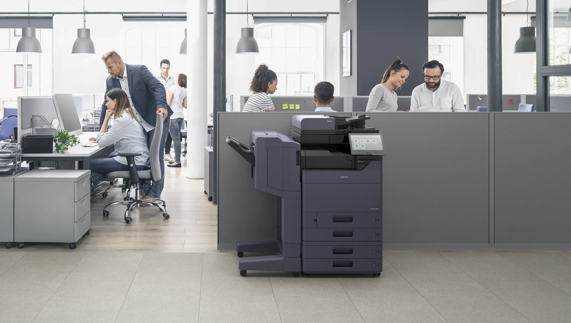 A Kyocera TASKalfa multifunction printer standing on the floor of a busy open plan office