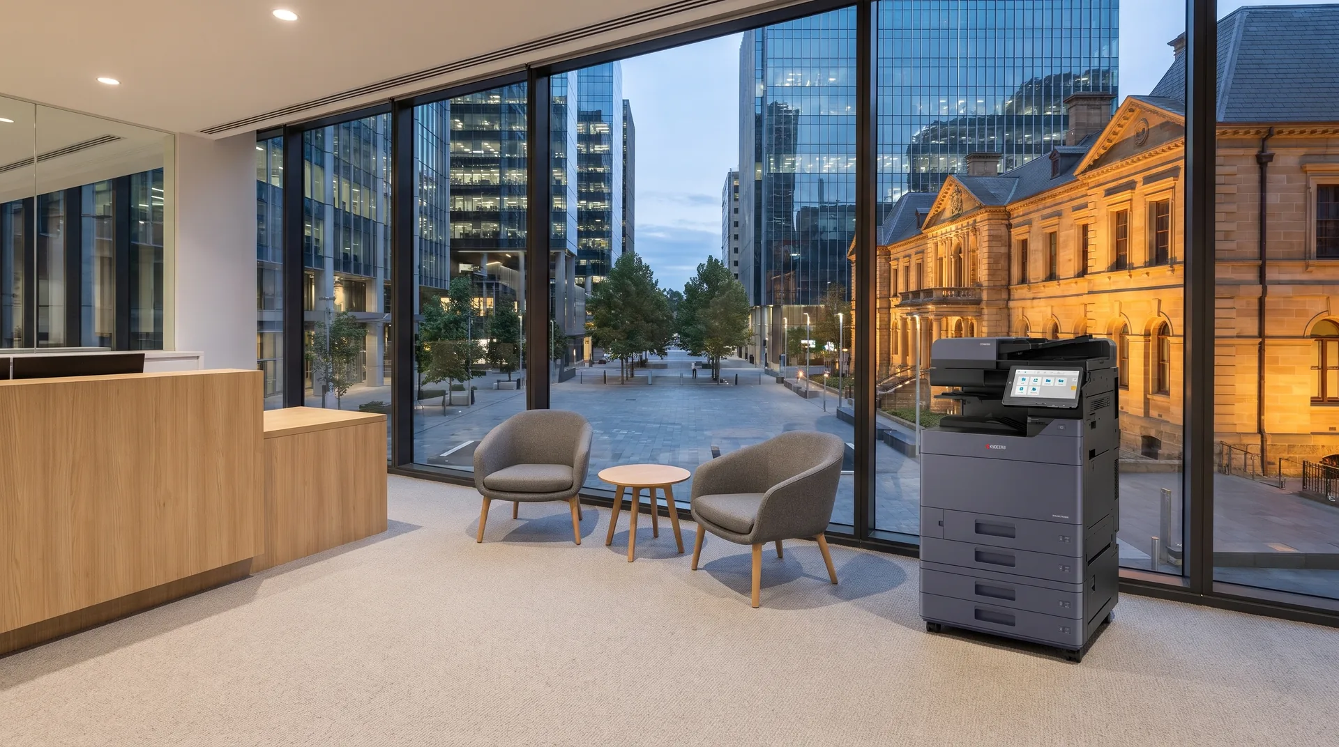 Kyocera photocopier on lease in a Parramatta reception area overlooking a city square with glass towers and a lit sandstone building at dusk
