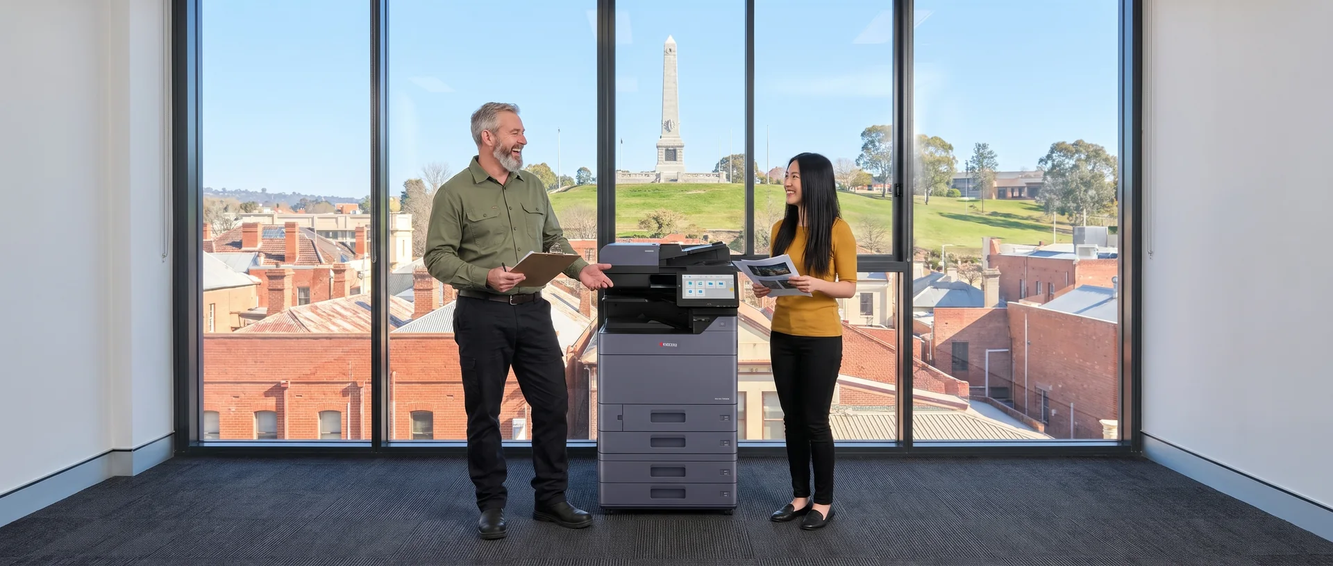 Bearded technician with a clipboard laughing with a young staff member beside a leased Kyocera copier, with Monument Hill in the window in Albury