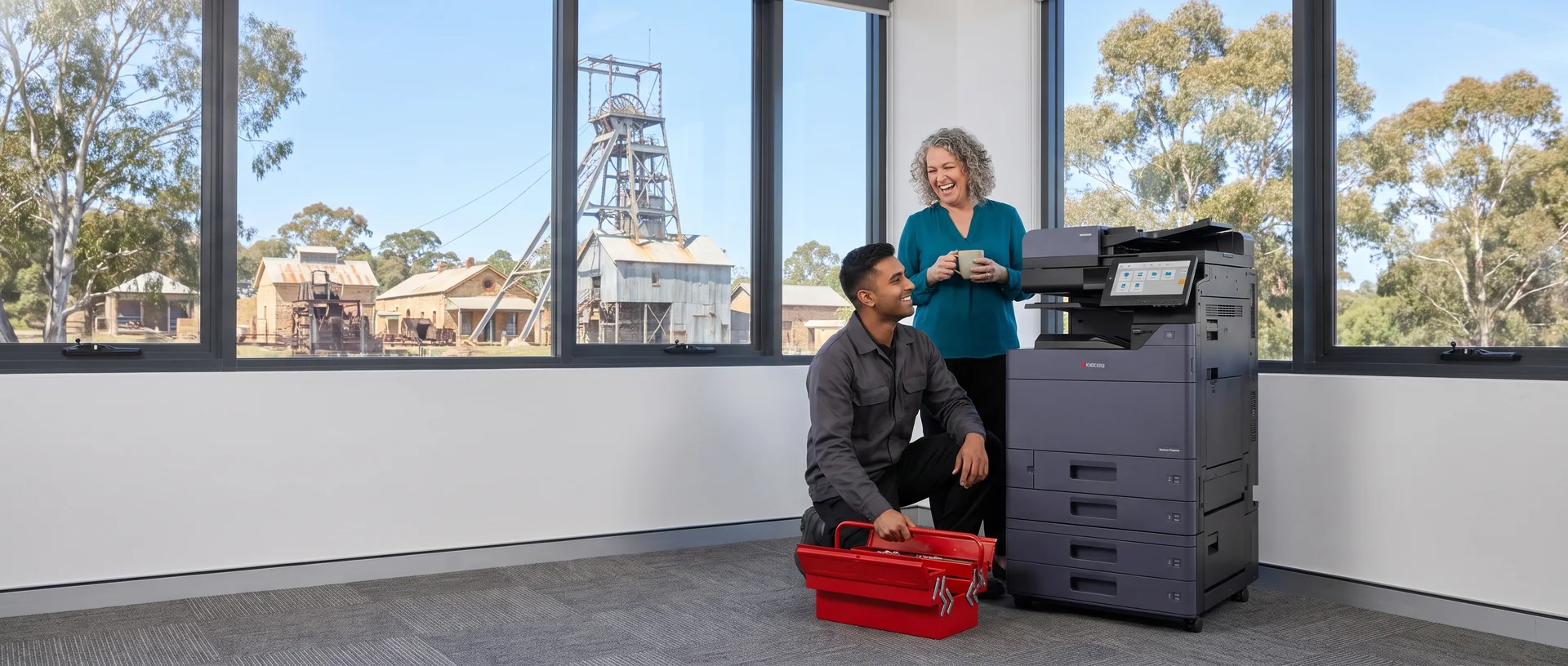 Technician kneeling by his toolbox beside a leased Kyocera copier, chatting with a smiling office manager, with the Central Deborah poppet head outside in Bendigo