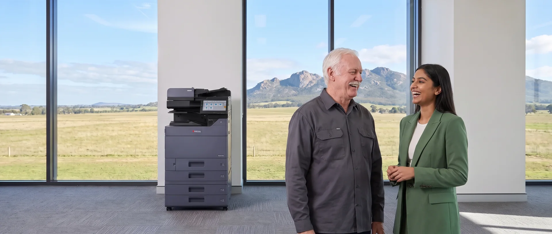 Technician laughing with a young office worker near a leased Kyocera printer in a Geelong office with the You Yangs outside