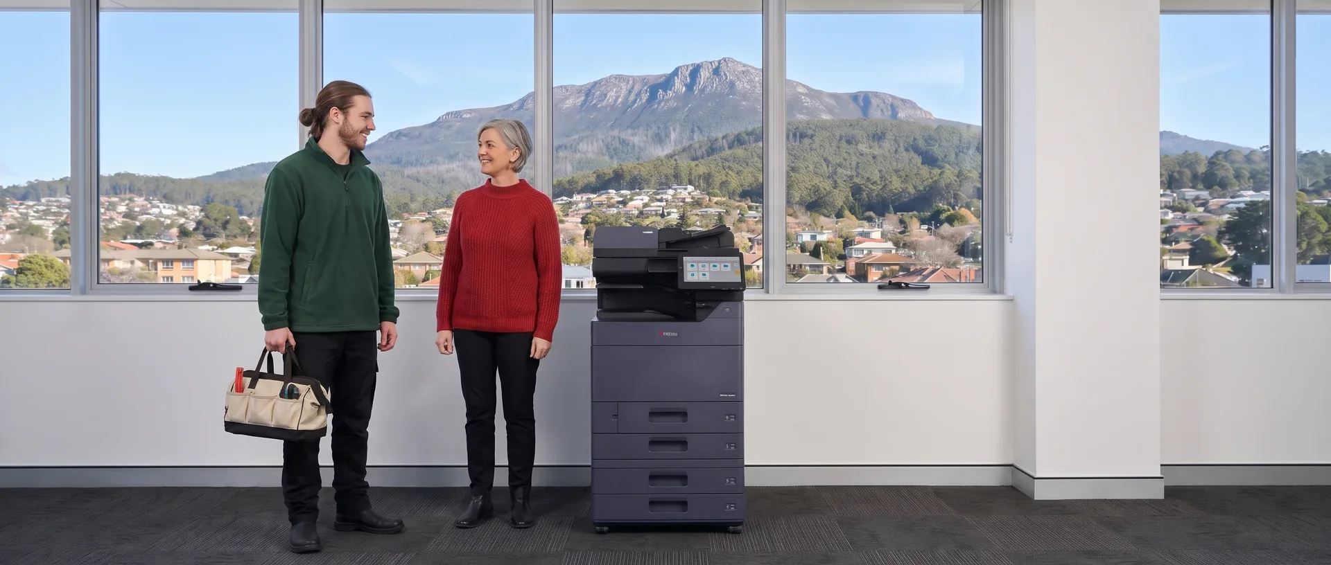 Technician with a tool bag chatting with an office manager beside a leased Kyocera printer in a Hobart office below kunanyi Mount Wellington